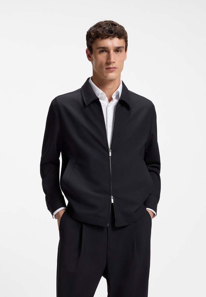 Jeune homme portant une veste noire zippée par-dessus une chemise blanche, les mains dans les poches, debout devant un fond blanc uni.