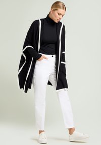 Vrouw die met één hand in de zak staat, gekleed in een zwarte coltrui, een zwart-witte gestreepte sjaal, witte broek en witte sneakers.