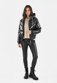 Veste bomber noire matelassée avec capuche, texture brillante, poignets élastiques et fermeture éclair à l'avant. Associée à des leggings noirs ajustés et des bottes noires imposantes.