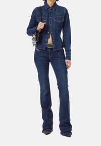 Chaqueta de mezclilla y vaqueros de campana en un tono oscuro, que presentan bolsillos en el pecho, costuras estructuradas y una hebilla de cinturón plateada. Los accesorios incluyen un bolso pequeño.
