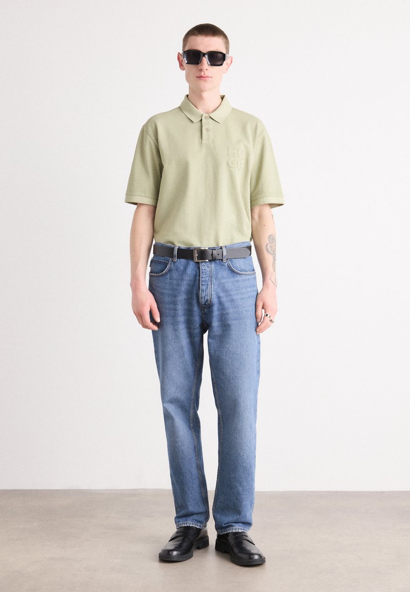 Polo en coton vert clair avec logo brodé, associé à un jean en denim bleu et une ceinture noire, complété par des lunettes de soleil noires et des chaussures noires.