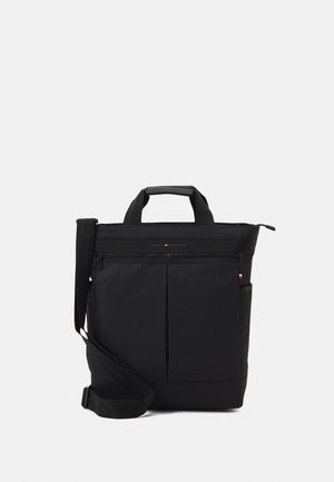 Sac fourre-tout noir vertical avec poignées courtes, poche zippée avant, poches latérales et bandoulière ajustable sur fond blanc.