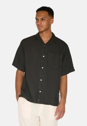 Short-sleeve black button-up shirt with a pocket, soft texture, and contrast stitching. Worn with light-colored pants.