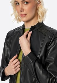 Black leather jacket featuring a stand-up collar, zip closure, and quilted shoulder accents. Worn over a green top, with fitted cuffs.