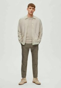 Cardigan bege claro por cima de um suéter a condizer, combinado com calças castanhas de xadrez e sapatos beges. Design simples, corte descontraído, texturas suaves.