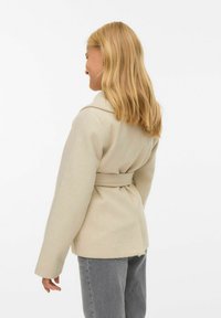 Bežinės vilnos mišinio paltas su ilgomis rankovėmis, plačiu apykakle, diržu juosmeniui ir struktūrizuotu siluetu, derinamas su pilkomis aukšto juosmens džinsais.