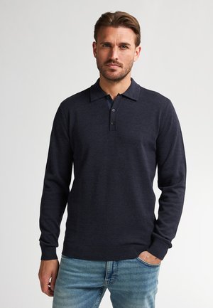 Marineblauw poloshirt met lange mouwen, een kraag, een placket met drie knopen en geribbelde manchetten. Draagt men bij lichte spijkerbroeken.