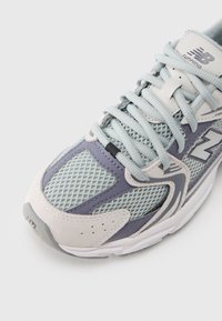 Light grey athletic shoe with mesh panels, purple accents, and textured suede. Features flat laces and a padded tongue for comfort.