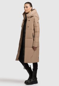 Lang beige puffer-frakke med hætte, lynlås og sidelommer. Har et quiltet design og sorte accenter, båret over sorte leggings og støvler.