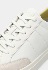Sneaker bianche in pelle con dettagli in suede grigio testurizzato, lacci piatti e finitura liscia. Presentano un design minimalista e un branding sottile.