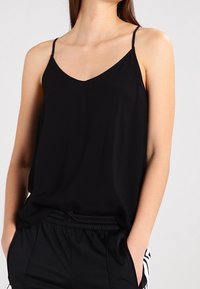 Donna che indossa un top nero a spaghetto infilato in pantaloni sportivi neri con strisce bianche sui lati, mani nelle tasche su uno sfondo semplice.