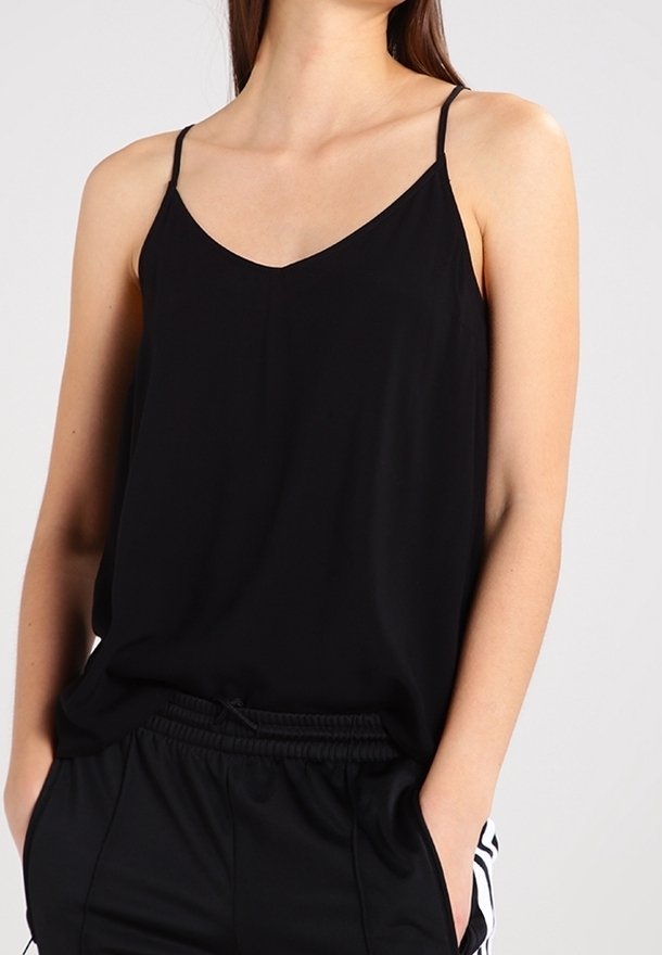 Donna che indossa un top nero a spaghetto infilato in pantaloni sportivi neri con strisce bianche sui lati, mani nelle tasche su uno sfondo semplice.