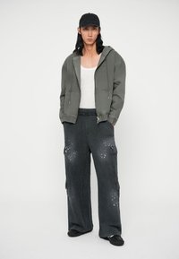 Sivá mikina s kapucňou a šnúrkami, biela tielko a tmavé cargo nohavice s vreckami a rozliatym detailom, skombinované s čiernym klobúkom a topánkami.