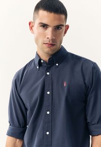 Marineblauw katoenen overhemd met knopen en opgerolde mouwen, met een klein roze kreeftenlogo op de linkerborst en witte knopen.