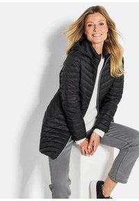 Zwarte pufferjas met een capuchon, die een doorgestikte uitstraling heeft en een ritssluiting aan de voorkant. Draagt over een witte trui en gecombineerd met grijze spijkerbroeken en zwarte sneakers.