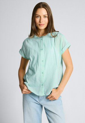 Jeune femme portant une chemise à manches courtes turquoise clair boutonnée et un jean bleu clair, debout les mains dans les poches devant un fond uni.