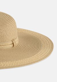 Szeroki kapelusz słomkowy w jasnym odcieniu brązu, z wplecioną fakturą i subtelnym plecionym akcentem wokół korony.