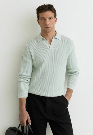 Mężczyzna o ciemnych włosach, ubrany w jasnozielony sweter z dzianiny i czarne spodnie, trzymający czarną torbę na jednolitym białym tle.