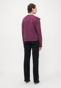 Donna in piedi di spalle che indossa una felpa a maniche lunghe viola, jeans neri a gamba dritta e scarpe con tacco nere su uno sfondo bianco.