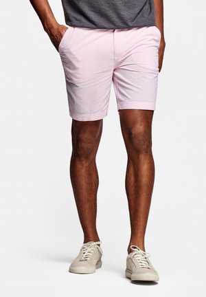 Homme portant un short rose clair, un t-shirt gris et des baskets beige, debout les mains dans les poches sur fond blanc.