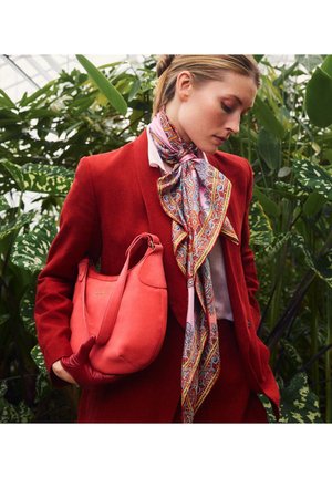 Vrouw in een rode jas en handschoenen houdt een koraalkleurige handtas vast, draagt een patroonrijke multicolour zijden sjaal en staat temidden van groene bladeren in een kassen.