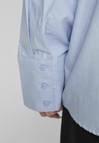 Camisa de manga larga de color azul claro con tres botones en los puños, textura suave y un corte ligeramente holgado con detalle de pliegue en los hombros.