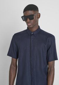 Korte mouwen marineblauwe shirt gemaakt van gestructureerde stof, met een button-down kraag en een enkele knopen sluiting. Zwarte oversized zonnebril.