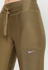 Olivově zelené sportovní legíny s hladkou texturou, stahovací pas a boční kapsa; obsahují stříbrné logo Nike na stehně.