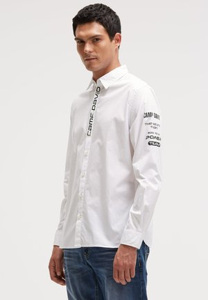 Mann trägt ein weißes Hemd mit Knöpfen, auf dem vorne und am Ärmel der Schriftzug "Camp David" zu sehen ist, kombiniert mit blauen Jeans, steht vor einem einfarbigen Hintergrund.