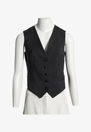 Chaleco negro sin mangas con botones y pequeños lunares blancos mostrado en un torso de maniquí blanco que lleva pantalones blancos.