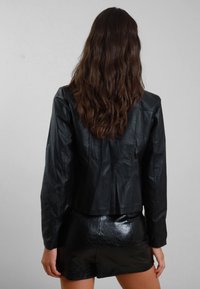 Giacca cropped in pelle nera con una vestibilità sartoriale e una texture liscia, abbinata a pantaloni corti neri lucidi. Vista da dietro, con i capelli che cadono.