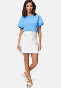 Blaues Baumwoll-T-Shirt mit Rüschensleeves, kombiniert mit einem weißen Jeansrock mit Vorderentaschen und silbernen Knöpfen. Weiße Sandalen zum Rutschen.