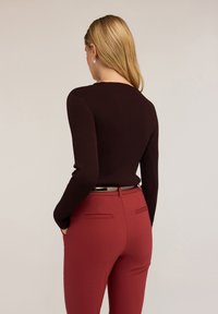 Donna con capelli biondi che indossa un maglione a coste marrone scuro e pantaloni rossi a vita alta, con le mani nelle tasche, di spalle.