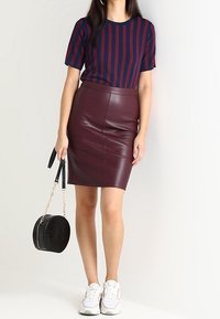 Femme portant un haut à rayures bleu marine et bourgogne à manches courtes, une jupe en cuir bourgogne, des baskets blanches, et tenant un sac à main rond noir.