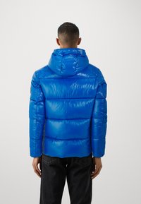 Mand iført en skinnende blå pufferjakke med hætte, set fra bagfra, parret med sorte jeans mod en ensfarvet baggrund.