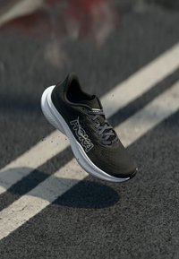 Chaussure de sport noire avec une tige texturée, semelle intermédiaire blanche et détail de logo. La chaussure semble être en mouvement au-dessus d'une surface au sol.