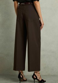 Wijdvallende, bruine broek met een gladde textuur, voorzien van twee achterzakken en een hoge taille. Gepaard met zwarte, open hakken.