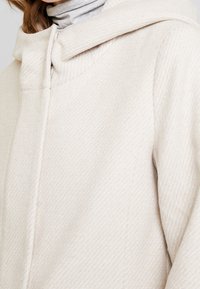 Ljust beige ullblandat ytterplagg med en texturerad yta, hög krage och knappstängning. Designen har en ren, minimalistisk silhuett.