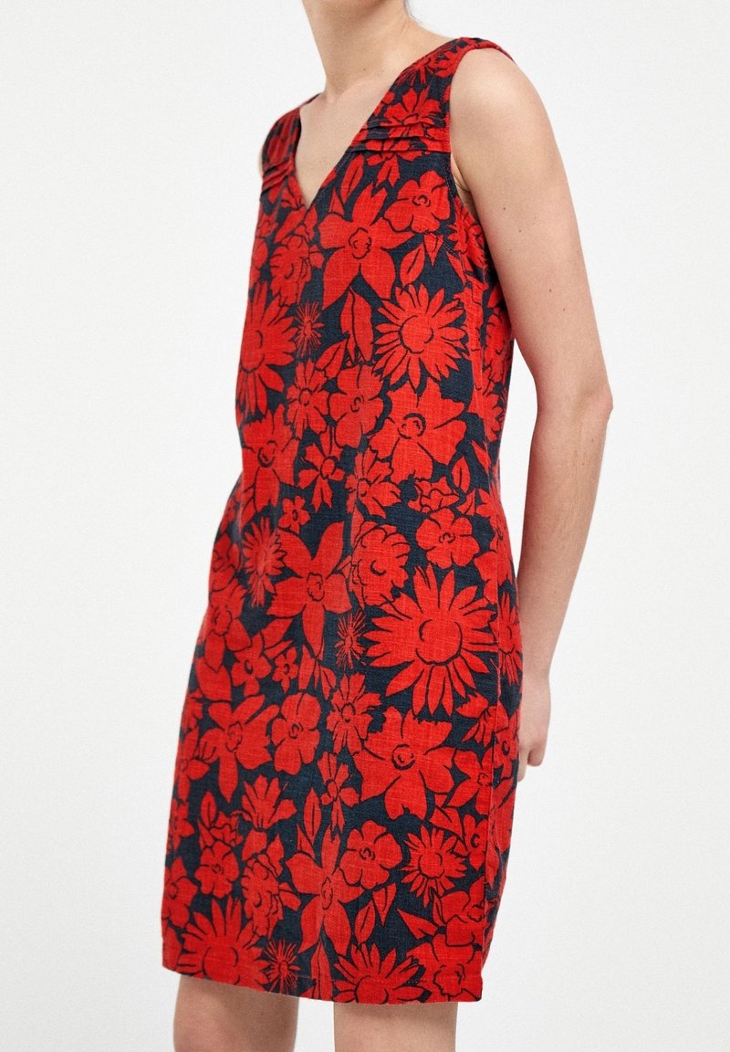 Robe rouge sans manches avec un grand motif floral noir, col en V, silhouette ajustée, portée par une personne sur un fond blanc uni.