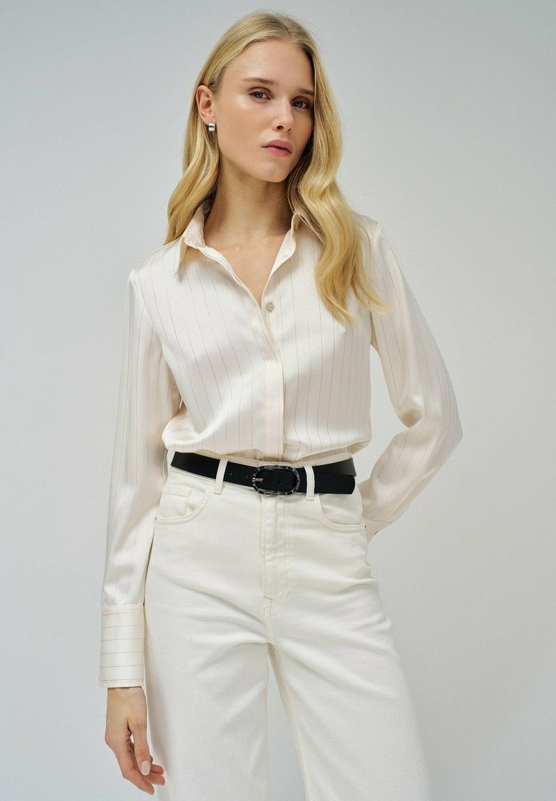 Blusa blanca a rayas con cuello de botones, combinada con jeans blancos de cintura alta y un cinturón negro con una hebilla circular.
