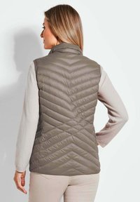 Gepolsterte graue Weste mit hohem Kragen, Zickzackmuster und leichtem Material, kombiniert mit einem hellen Pullover und beigen Hosen.