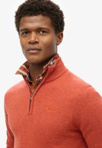 Maglione rosso con zip e collo a coste. Sotto, una camicia a scacchi nei toni dell'arancione, verde e blu. Focus sulla trama e sui dettagli del lavorato a maglia.