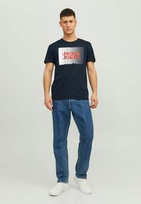 Tricou din bumbac bleu marin, cu un grafic dreptunghiular care afișează textul "JACK & JONES" în dungi roșii și albe. Asortat cu blugi albaștri și adidași albi.