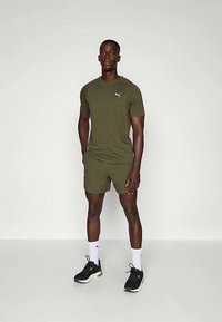 Olivgrönt träningsset bestående av en figursydd kortärmad tröja och shorts, tillverkat av fukttransporterande material, kombinerat med svarta sneakers.