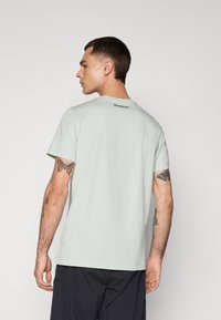 Man facing away wearing a pale green Mammut t-shirt and black pants, showing tattoos on both upper arms against a plain background.