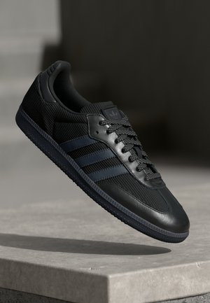 Czarny sneaker Adidas z panelami z siateczki i skóry, trzema ciemnymi paskami po boku, wzorzystymi sznurowadłami i gumową podeszwą zawieszoną nad powierzchnią betonową.