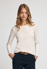 Cremfarbens Langarmshirt aus weichem Stoff, mit Rundhalsausschnitt und kleinem gesticktem Logo. Kombiniert mit dunklen Hosen und einem braunen Gürtel.