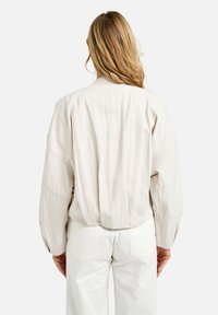 Light beige bomber jacket with ribbed collar, dropped shoulders, and textured fabric. Paired with white pants, showing minimalistic design.