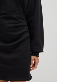 Schwarzes Langarmkleid mit seitlichen Raffung und texturiertem Finish. Bietet einen lockeren Schnitt und gerippte Bündchen. Minimalistisches Design ohne Muster.