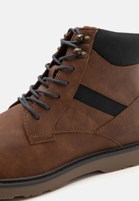 Bota de tobillo de cuero marrón con acentos de textil negro, que presenta cordones grises, ojales de metal y una suela texturizada.